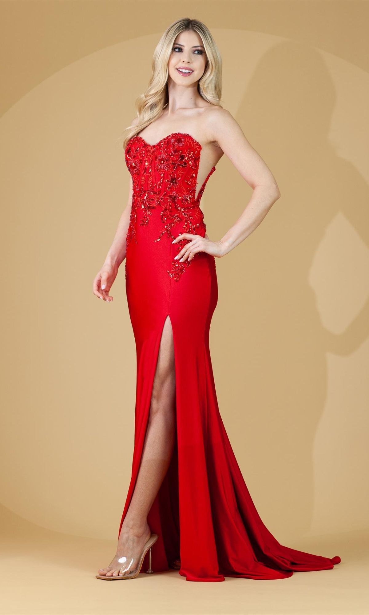 A woman in the Strapless Sweetheart Sheer-Sides Long Prom Dress 7052, featuring a thigh-high slit and beaded details, stands in sheer heels against a beige background.
